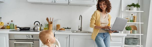 niña mirando a la madre alegre de pie con el ordenador portátil en la cocina, equilibrio de la vida laboral, pancarta - Foto, imagen