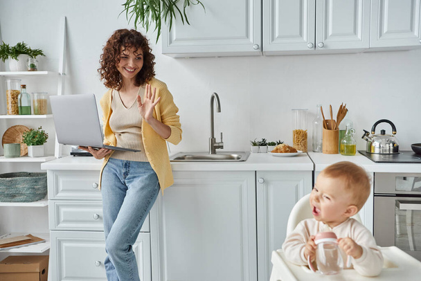 радостная женщина с ноутбуком размахивая рукой маленькой дочери с бутылочкой ребенка на кухне, внештатная работа - Фото, изображение