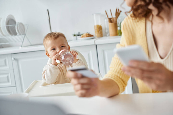 dziewczynka pije z butelki dla niemowląt w pobliżu mamy ze smartfonem i kartą kredytową na niewyraźnym pierwszym planie - Zdjęcie, obraz