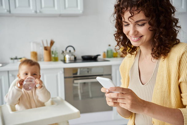 Улыбающаяся женщина отправляет сообщения по мобильному телефону, пока маленький ребенок пьет из детской бутылочки на кухне - Фото, изображение