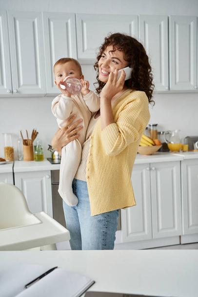vrolijk moeder praten op smartphone en houden kind drinken uit baby fles in de keuken - Foto, afbeelding