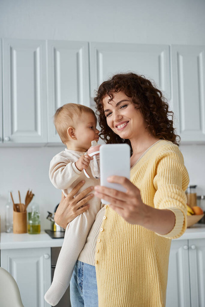 örömteli nő csinál szelfi a mobiltelefon kisgyermek lánya gazdaság cumisüveg a konyhában - Fotó, kép