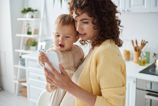 mujer sosteniendo niño emocionado y navegar por Internet en el teléfono inteligente en la cocina, crianza moderna - Foto, imagen