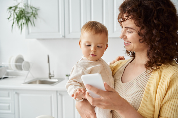 cheerful woman showing mobile phone to toddler girl while standing in modern kitchen, digital age - Photo, Image