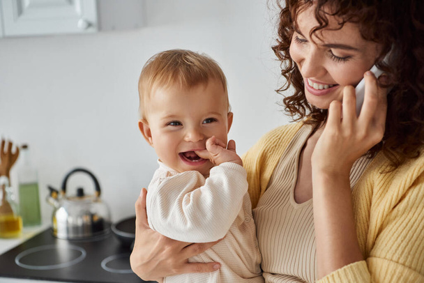 улыбающаяся женщина, держа весёлую дочку и разговаривая по смартфону на кухне, счастье - Фото, изображение