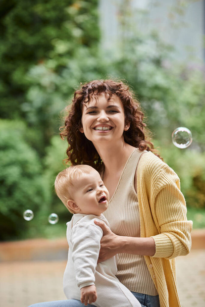 屋外の空飛ぶ石鹸の泡の近くに赤ん坊の娘との興奮した女性の肖像画,幸せな瞬間 - 写真・画像