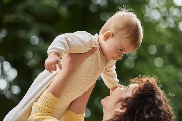 mujer feliz jugando con la hija del bebé y criándola en manos al aire libre, maternidad dichosa - Foto, imagen
