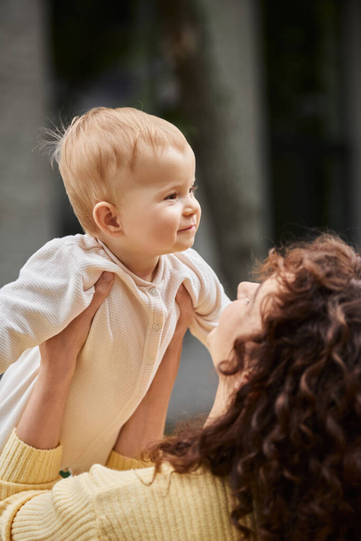 woman with wavy hair holding pleased little daughter in romper while spending time outdoors - Photo, Image