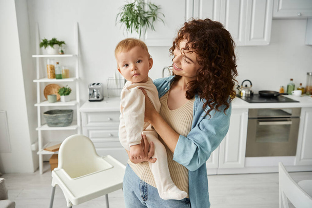 fröhliche Frau hält entzückende kleine Tochter im Strampler, während sie in der modernen Küche steht - Foto, Bild