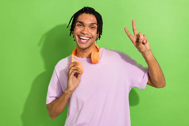Photo of funky positive man beaming smile touch hang neck headphones demonstrate v-sign isolated on green color background. - Φωτογραφία, εικόνα
