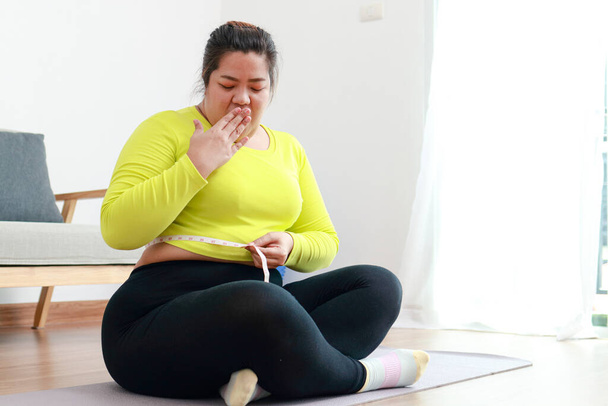 Mujer asiática gorda haciendo ejercicio en casa usando una cinta métrica para medir su circunferencia de cintura. Parece sorprendida por el tamaño. Concepto deportivo, atención médica. pérdida de peso - Foto, Imagen