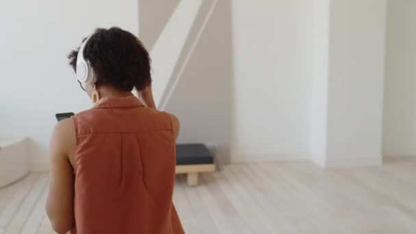 Mittlere Folgeaufnahme einer jungen Frau mit Brille, Kopfhörer und Smartphone, die durch eine stilvolle minimalistische Wohnung läuft, sich auf die Couch setzt, zur Musik schaukelt und lächelt - Filmmaterial, Video