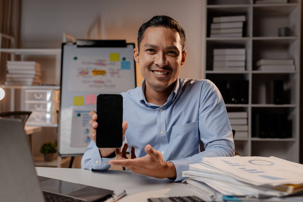 Ázsiai férfi mobilt tart egy startup cég irodájában, a cég alkalmazottja túlórázik, késő esti túlórázik egy elfoglalt és keményen dolgozó céges alkalmazottal. A túlóra fogalma. - Fotó, kép