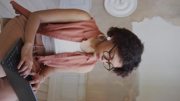 Μέση κάθετη λήψη νεαρής Αφροαμερικανής γυναίκας με κοντά καστανά κυματιστά μαλλιά, σε γυαλιά που κάθονται σε σκαμνί στο σπίτι και πληκτρολογούν σε φορητό υπολογιστή, σε άδειο δωμάτιο - Πλάνα, βίντεο