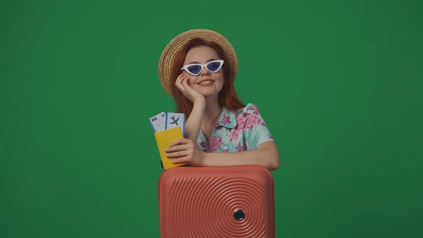 Travelling advertisement creative concept. Woman traveller in straw hat and glasses with suitcase holding flight tickets, smiling looking up and dreaming. Isolated on green background. - Photo, image