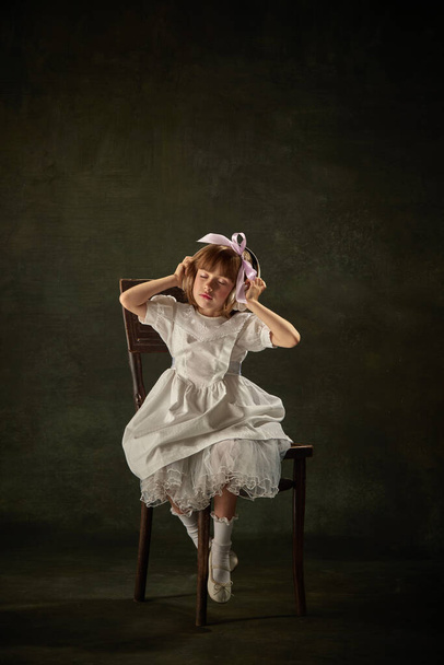 Porträt eines ruhigen kleinen Mädchens, das mit Kopfhörern Musik hört und auf einem Stuhl vor Vintage-Hintergrund sitzt. Märchenprinzessinnenfigur des Märchens. Konzept der Fantasie, Studie, Zukunft, Gadgets, Studium. Anzeige - Foto, Bild
