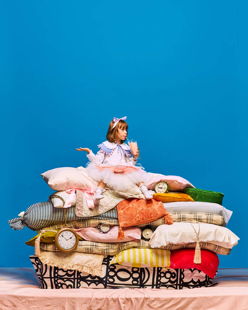 Portrait of little princess sitting on big pile of pillows drinking fresh, healthy cocktail over blue background. Fairy character eating breakfast. Concept of fairy remake, healthy lifestyle, food. - Photo, Image