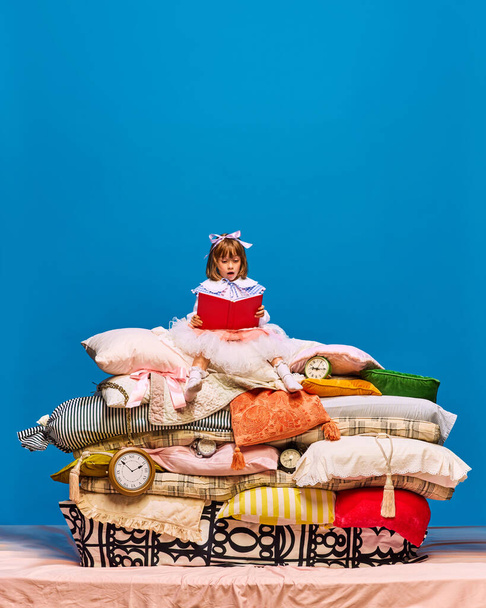 Портрет маленької принцеси, що сидить на великій купі подушок, читає книгу в червоній обкладинці на синьому фоні. Казковий персонаж вчиться. Поняття казкового рімейку, здорового способу життя, книжкового тепла, книгарні. Реклама - Фото, зображення