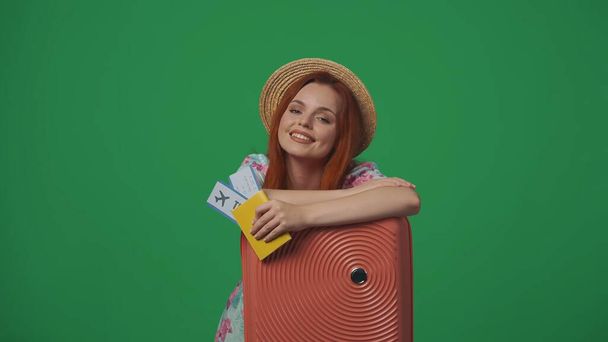 Travelling advertisement creative concept. Woman traveller in straw hat holding flight tickets, hand on suitcase, smiling happy face expression. Isolated on green background. - Foto, Bild
