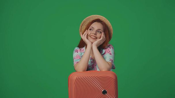 Travelling advertisement creative concept. Woman traveller in straw hat with happy smiling face expression dreaming, hands on suitcase. Isolated on green background. - Photo, image
