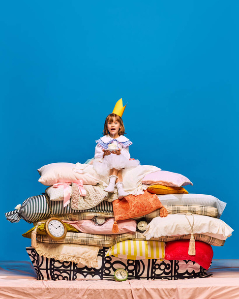 Retrato de surpresa, pequena princesa feliz em coroa amarela sentado em grande pilha de almofadas com doce, saboroso, delicioso bolo. Conceito de remake de fadas, comida, bebidas, feriados, sobremesas. Anúncio - Foto, Imagem