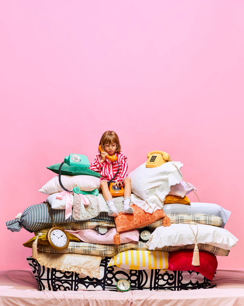 Retrato de la pequeña princesa bonita sentada en la cama de plumas grandes con almohadas hablando en teléfonos retro sobre fondo de estudio rosa. Concepto de negocios, remake de hadas, infancia, conversación. Anuncio - Foto, Imagen