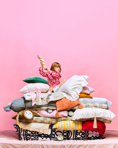 Ritratto di principessa arrabbiata seduta su un grande letto di piume con cuscini con baguette francese su sfondo rosa da studio. Concetto di remake delle fate, mattina, cibo tradizionale, bevande. Pubblicità - Foto, immagini