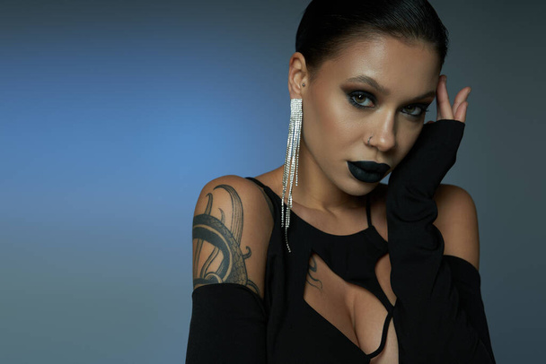 sinister beauty, tattooed woman in dark makeup and black halloween dress on blue and grey backdrop - Photo, Image