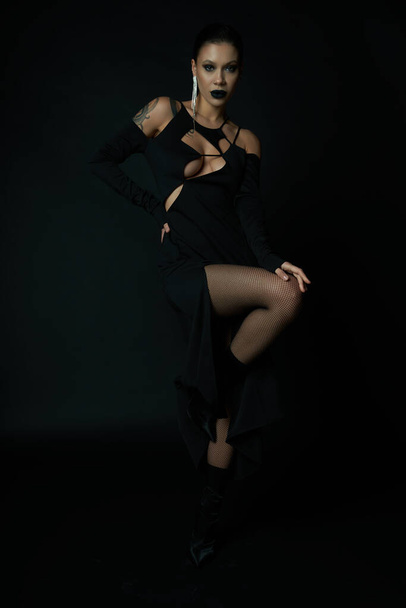 σαγηνευτική γυναίκα σε σκούρο μακιγιάζ και κομψό αποκριάτικο φόρεμα κοιτάζοντας κάμερα σε μαύρο, γοτθική ομορφιά - Φωτογραφία, εικόνα