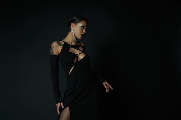 sensualité gothique, femme tatouée en robe enchanteresse sexy et maquillage sombre regardant loin sur noir - Photo, image
