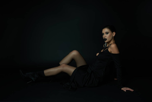 femme élégante en costume de sorcière sombre assise et regardant la caméra sur noir, beauté sinistre - Photo, image