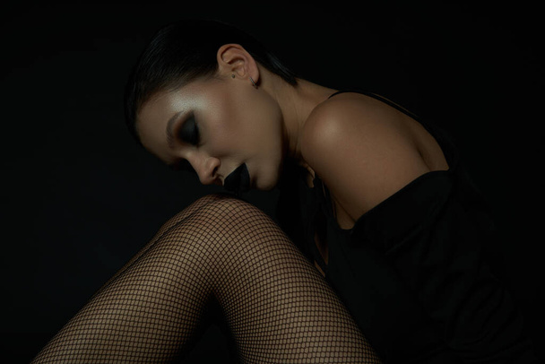 gothic glamour, stylish woman with closed eyes and dark makeup sitting in fishnet tights on black - Photo, Image