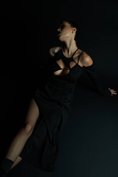 παθιασμένη γυναίκα με τατουάζ στο σέξι αποκριάτικο φόρεμα κάθεται σε μαύρο φόντο, γοτθική ομορφιά - Φωτογραφία, εικόνα