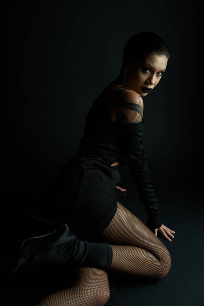 mujer apasionada en vestido sexy y medias de rejilla sentado y mirando a la cámara en el fondo negro - Foto, Imagen