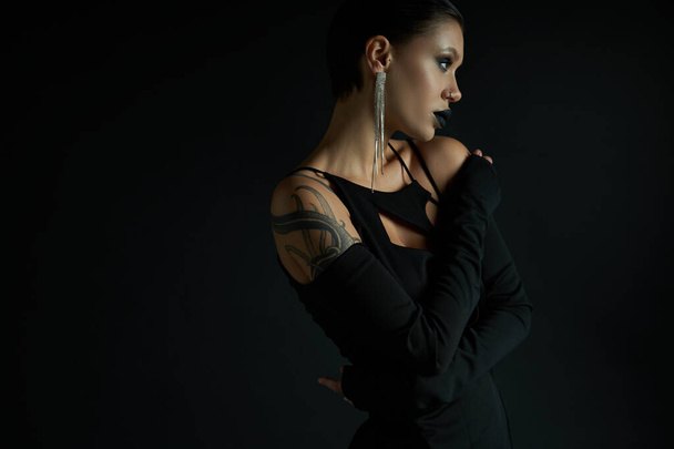 tattooed and glamour woman with eerie makeup posing in sexy dress on black, halloween concept - Foto, imagen