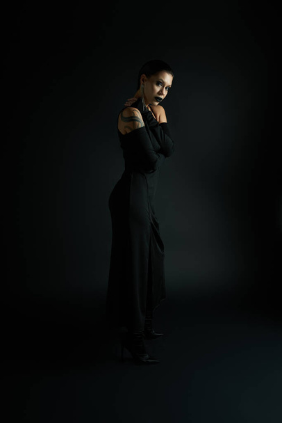 sexy mujer tatuada con labios oscuros usando elegante vestido de halloween y mirando a la cámara en negro - Foto, imagen