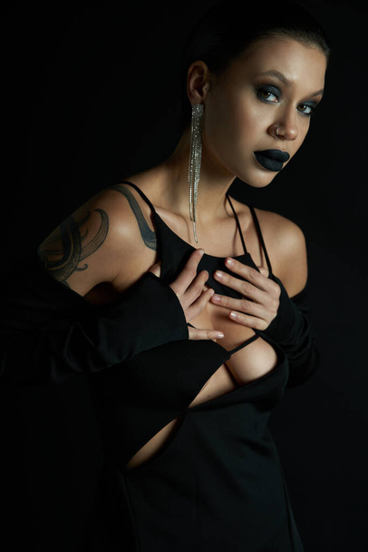 γοτθική γοητεία, γυναίκα με τατουάζ σε σέξι αποκριές φόρεμα αγγίζοντας το στήθος και κοιτάζοντας κάμερα σε μαύρο - Φωτογραφία, εικόνα