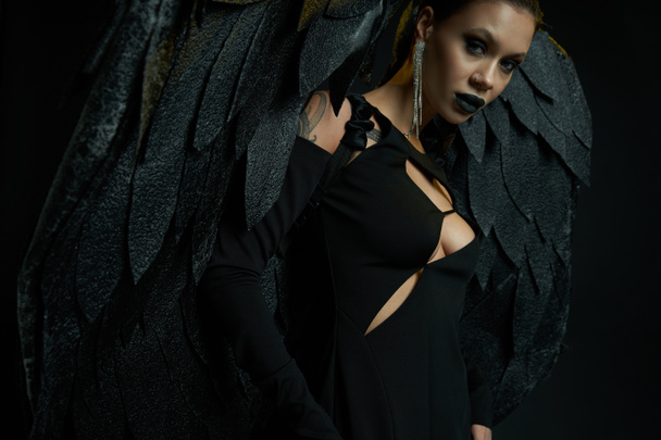σαγηνευτική γυναίκα με τατουάζ στο αποκριάτικο κοστούμι του σκοτεινού δαίμονα με φτερά κοιτάζοντας κάμερα στο μαύρο - Φωτογραφία, εικόνα