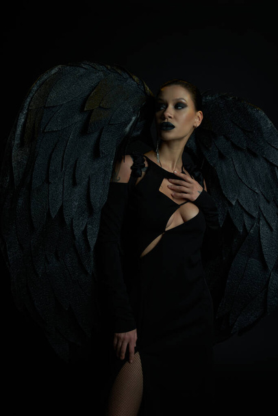 tattooed woman in halloween costume of fallen angel with wings looking away on black, demonic charm - Photo, Image