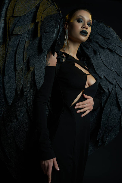 dark beauty, tattooed woman in halloween costume of winged fallen angel looking at camera on black - Photo, Image