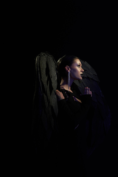 side view of woman in costume of fallen angel with dark wings praying in darkness, black backdrop - Photo, Image