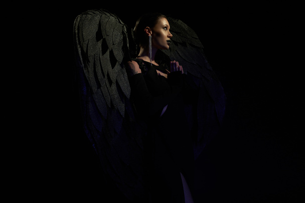 vista lateral de mujer misteriosa disfrazada de criatura alada demoníaca rezando sobre fondo negro - Foto, imagen