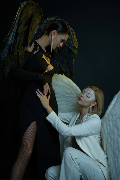 women in Halloween costumes, white angel sitting on haunches near dark demon on black backdrop - Photo, Image