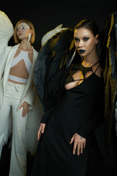 démonická žena dívá na kameru poblíž anděla na černém pozadí, ženy v kostýmech okřídlených tvorů - Fotografie, Obrázek
