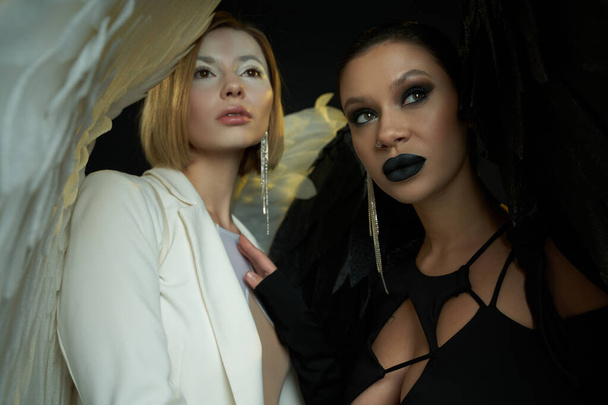 mujeres en disfraces de Halloween de ángel y demonio mirando hacia otro lado en el fondo negro, la guerra espiritual - Foto, imagen