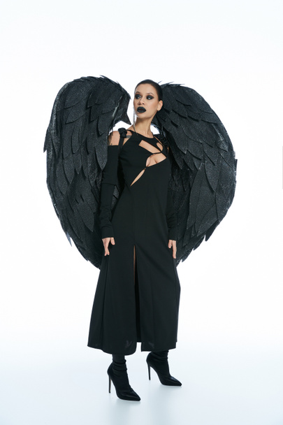 in voller Länge geheimnisvolle Frau im Kostüm eines schwarzen geflügelten Dämons, der vor weißem Hintergrund wegschaut - Foto, Bild