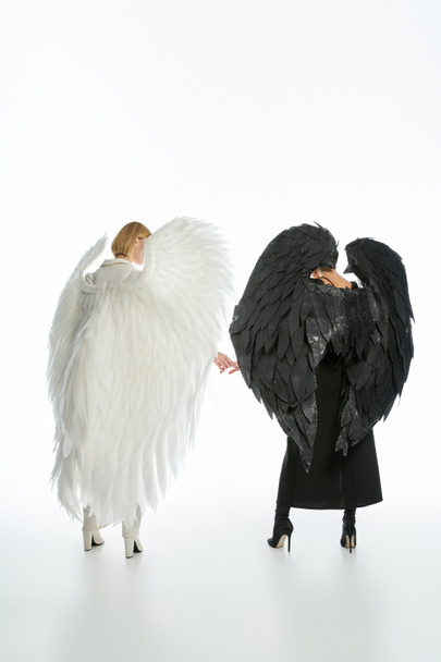 vista posterior de las mujeres en trajes de diablo y ángel con alas negras y ligeras cogidas de la mano en blanco - Foto, imagen