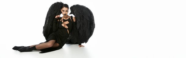 mujer sexy en maquillaje oscuro y traje negro de demonio alado sentado en blanco, bandera - Foto, Imagen