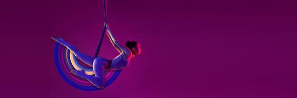 Młoda, elastyczna dziewczyna uprawiająca jogę powietrzną, ćwicząca na fioletowym tle w neonowym świetle z abstrakcyjnymi elementami designu. Kolaż sztuki współczesnej. Pojęcie sportu, kreatywności, działania i ruchu, zdrowia - Zdjęcie, obraz