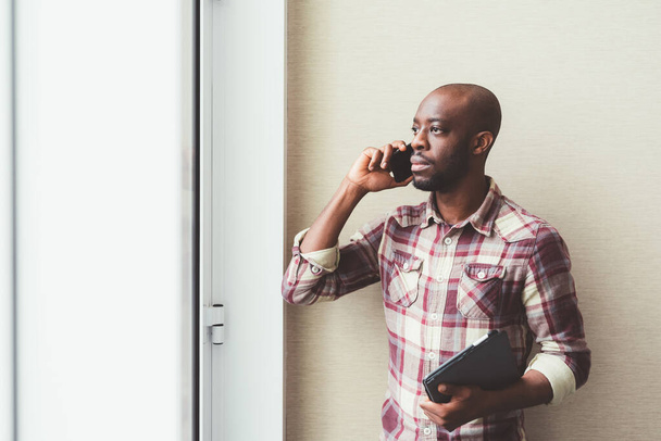 černý muž sedí blízko okna a mluví smartphone  - Fotografie, Obrázek
